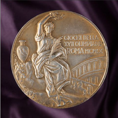 Yellow-coloured circular medal with a classical image showing a woman dressed in a robe, sitting cradling a small branch in her right arm and holding a laurel above her head with her left. An urn apears to her left and part of an arched bridge to her right. The medal has been inscribed with the text 'GIOCHI DELLA / XVII OLIMPIADE / ROMA MCMLX'