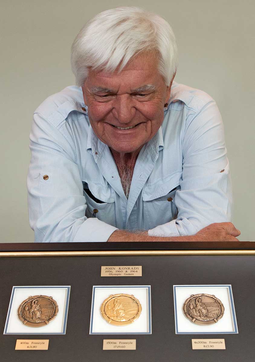 Colour photograph of a man resting with folded arms on a table. In front is a framed set of three circular medals.