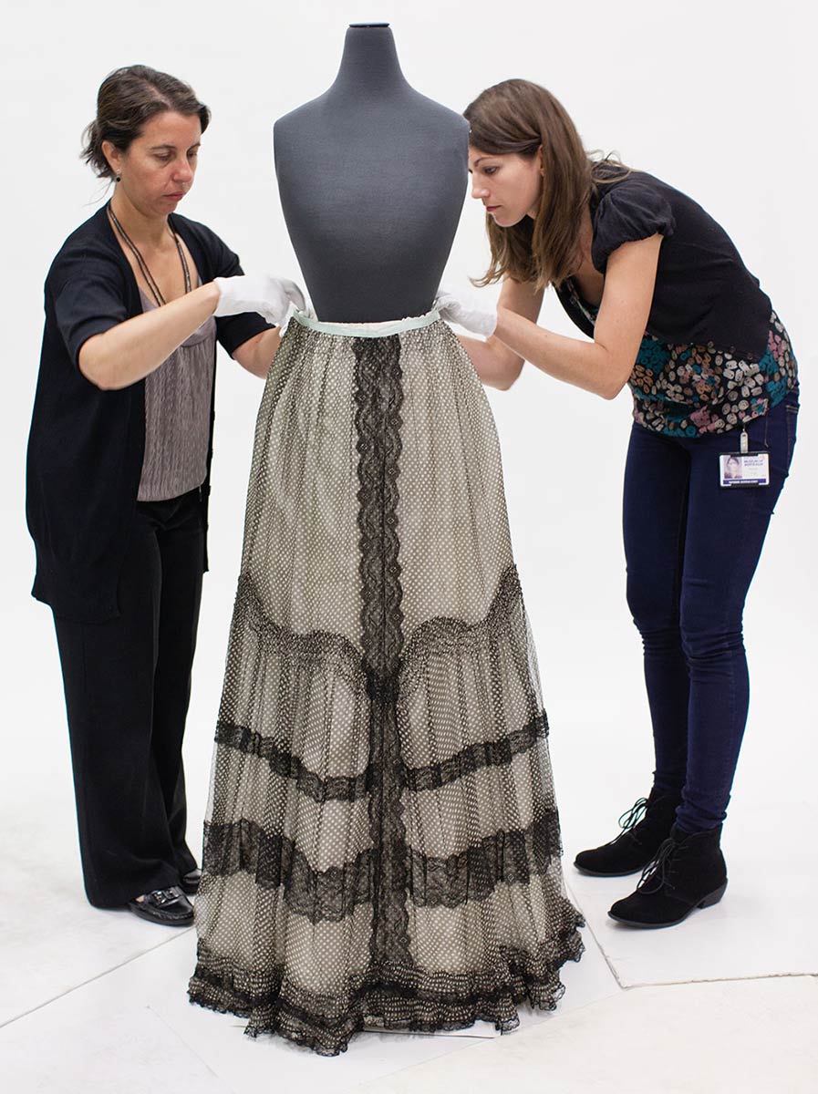 Two women stand either side and slightly to the rear of a mannequin. The mannequin's torso has neither arms nor a head and is finished in a dark grey material. The women, both wearing short white gloves, lean in to adjust the waistband of a skirt, which covers the lower half of the mannequin. The long skirt is mainly light grey in colour, with black lace down the centre, around the hem and in three vertical bands from about knee to hem.