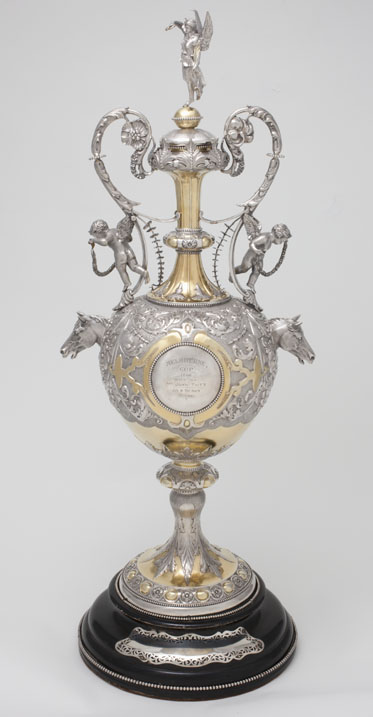A silver-gilt chased cup with gold leaf, consisting of a base, cup and lid. The cup is decorated with silver foliage designs and two silver horse heads on either side. A central shield is inscribed with the words ''MELBOURNE / CUP / 1866 / WON BY / MR JOHN TAIT'S / BLK. H. The Barb / 3 years'. Two cherubs are attached to the ornate handles at the top of neck, and an angel holding a wreath sits on the top of the lid. The circular base is painted black.