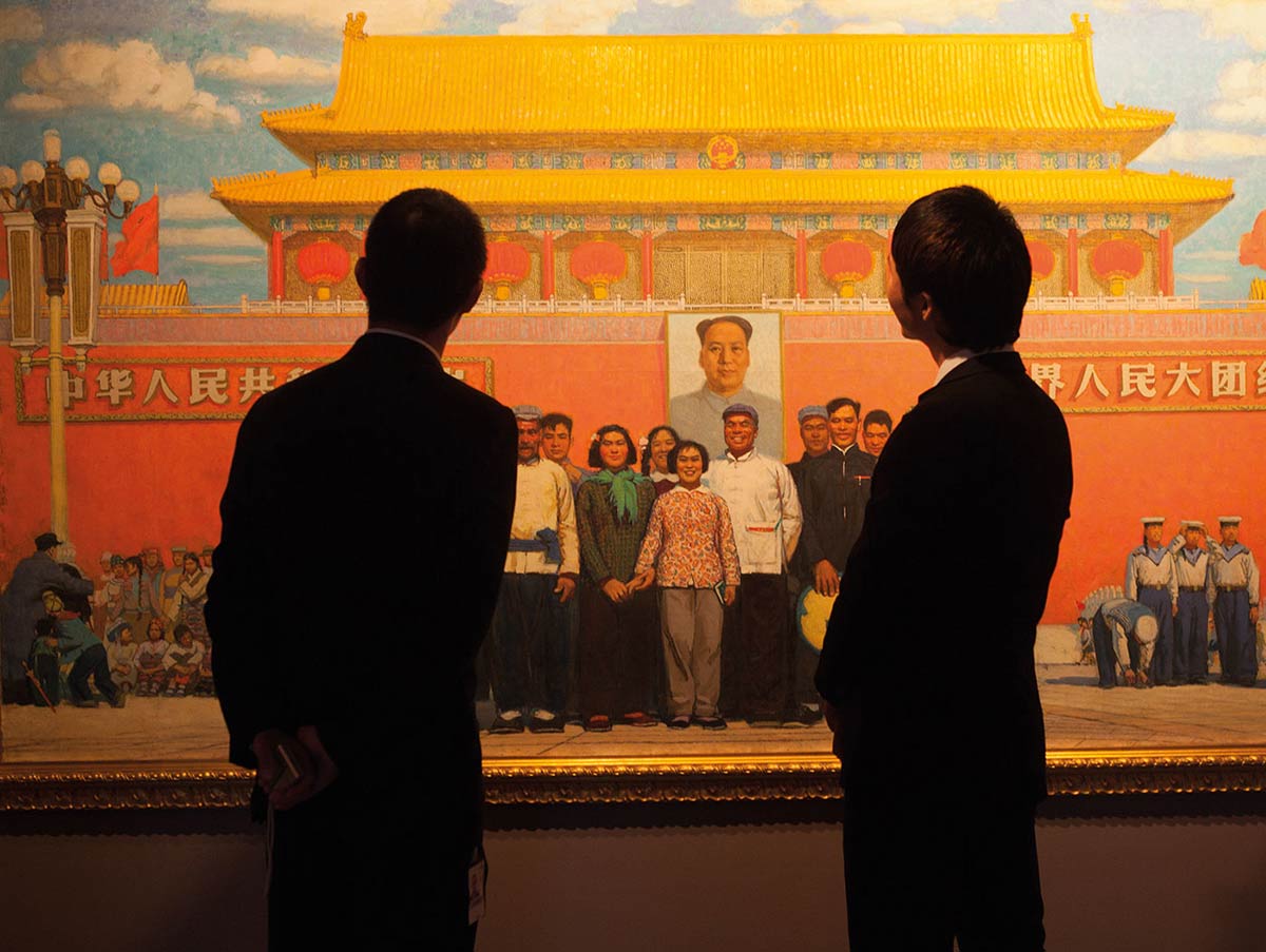 A colour image of two people dressed in dark suits viewing a large painting mounted on a wall.