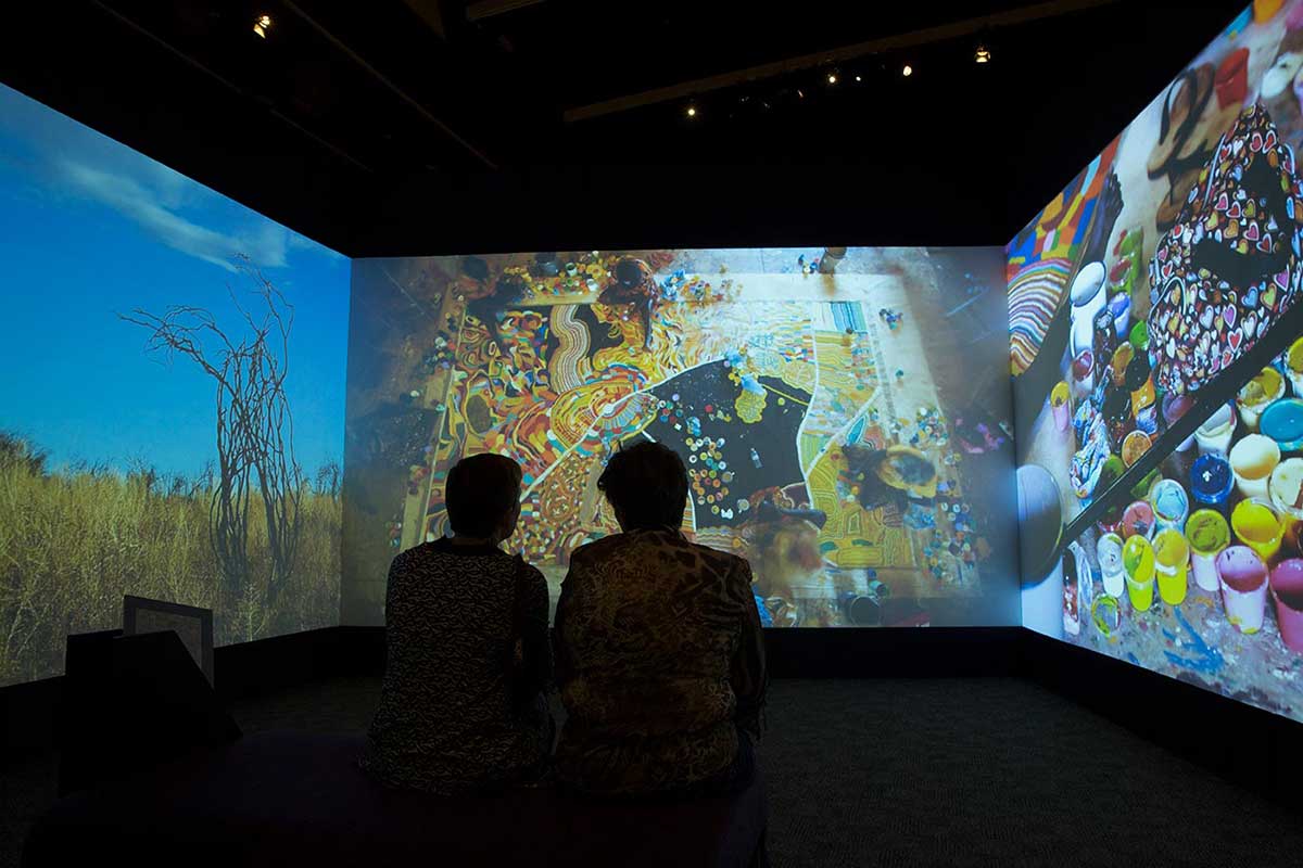 Silhouettes of two seated people watch a large and colourful video installation.