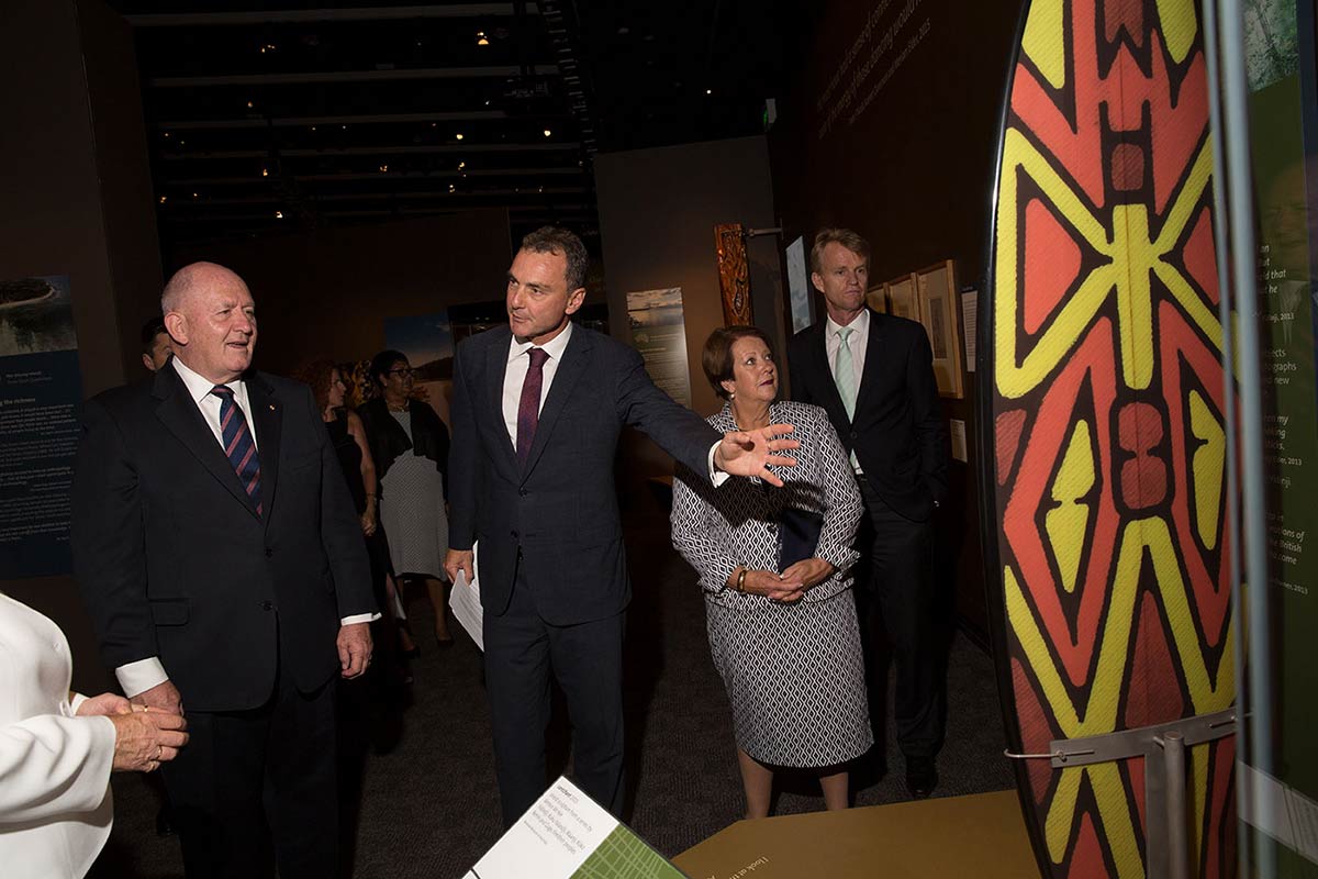 Three men and a woman in an exhibition look at shield painted with red and yellow geometric symbols.