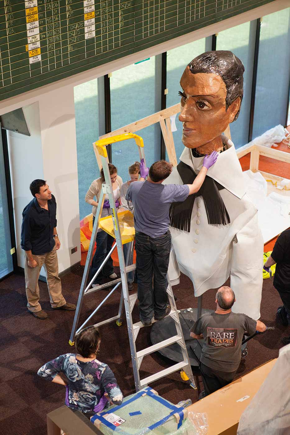 Photograph taken from above, looking down at an oversized puppet consisting of a female face, constructed of painted papier-mache. The woman wears a white collared-shirt and black taselled scarf. A man on the third rung of a large ladder reaches up to adjust the puppet's collar. Five other people stand in the gallery space.