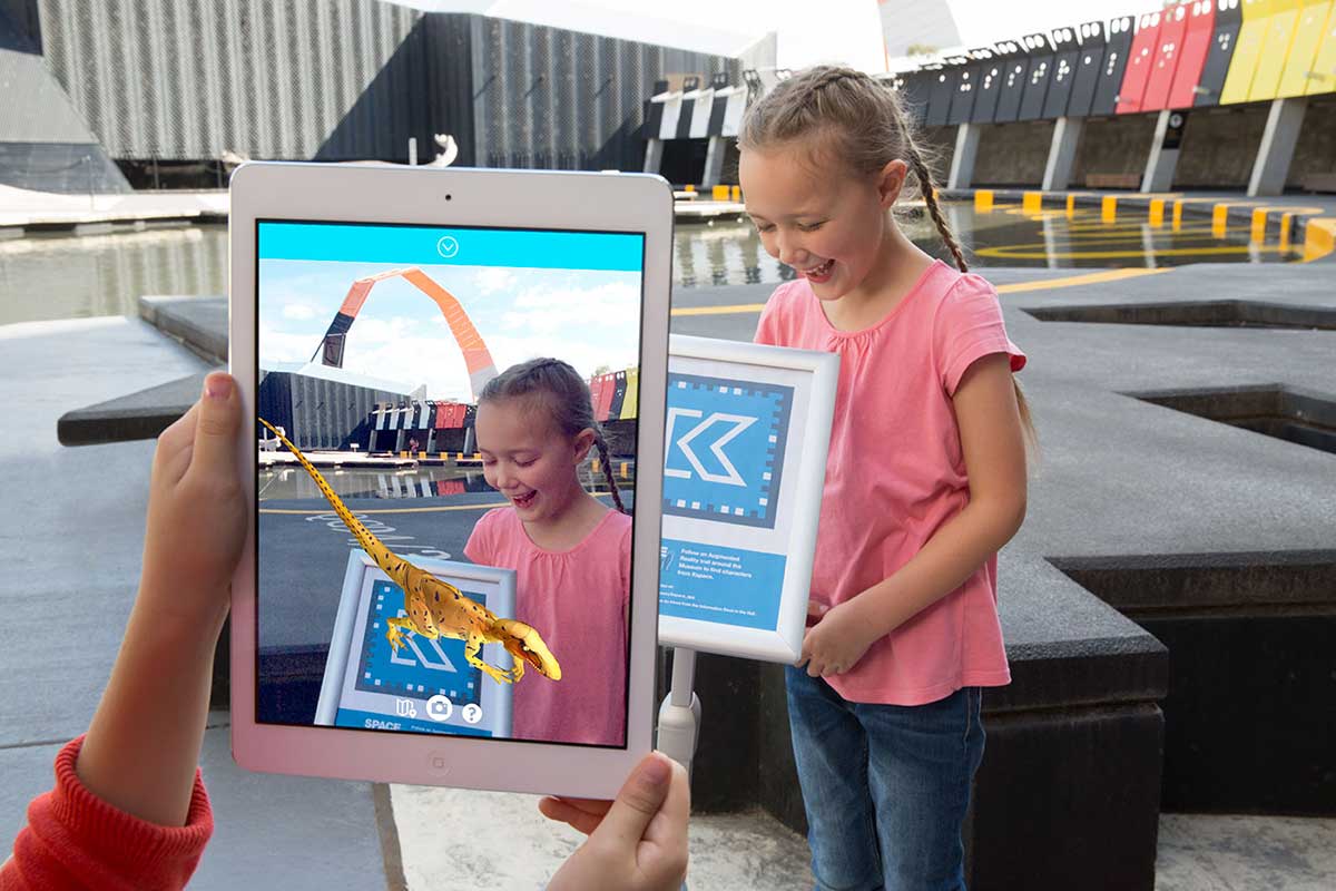 A young girl uses an electronic tablet to play a game which shows a virtual dinosaur.