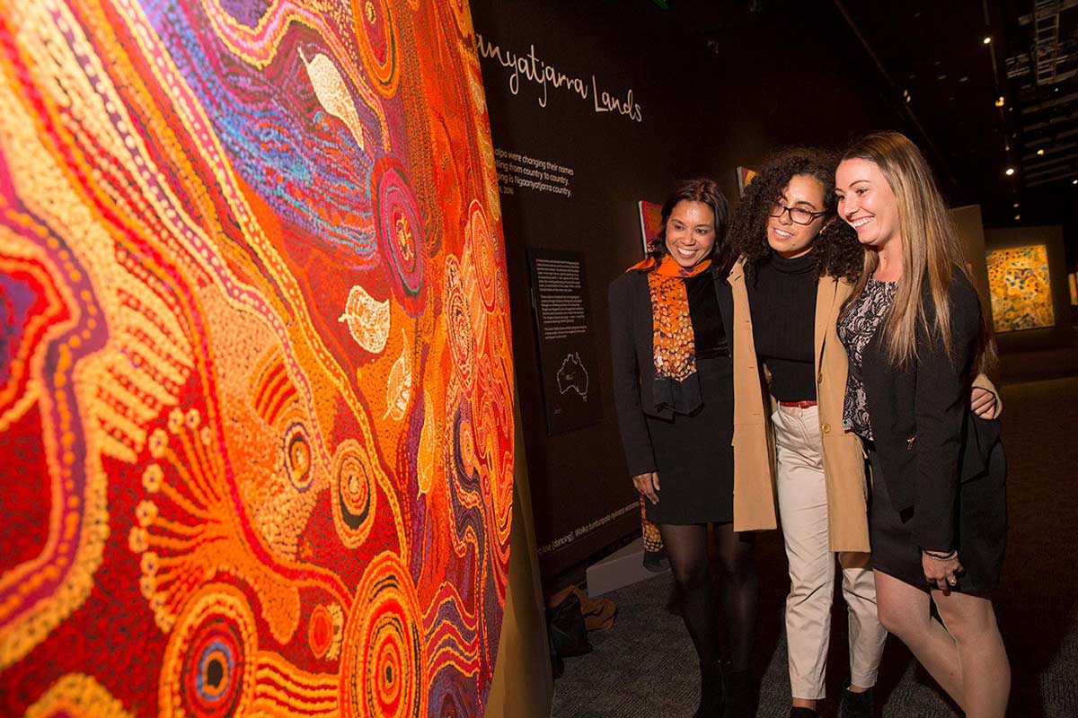 Three women admire a circular painting in the 'Songlines' exhibition.