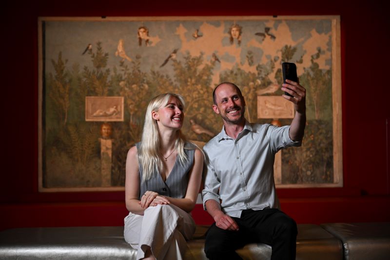 A man and a woman sit on a bench in an exhibition space and take a selfie with a fresco behind them