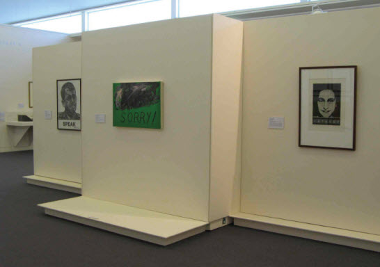 Installation shot showing three works, a portrait with the word 'Speak' at the bottom, a painting with the word 'Sorry!' in the lower right corner and a portrait with words at the bottom that are too small to read.