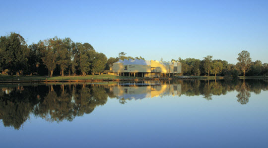 View of the Benalla Art Gallery taken from across a river. The gallery is a modern building in green colours. The top third of the image is clear blue sky, the bottom half shows river and the gallery's reflection in the water.
